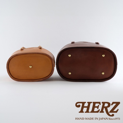 職人手作皮革束口手提包，左為淺棕色，右為深棕色，兩包均為橢圓形底部，配有四個黃銅腳釘，底部中央亦有一個黃銅釘。