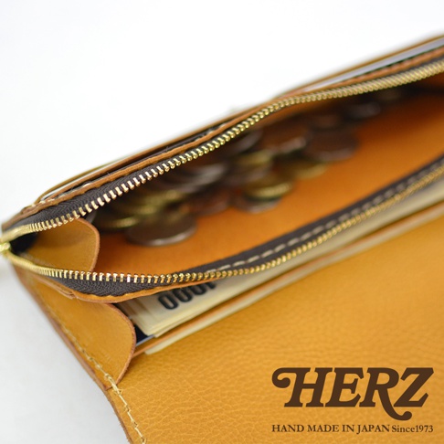 職人手作皮革長夾，特寫打開的拉鍊零錢袋，內有硬幣，並可見部分紙鈔。整體為芥末黃色皮革，搭配深棕色拉鍊與金色拉頭，車縫線細緻。