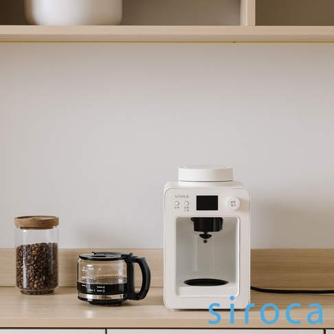 白色 Siroca SC-A3520 自動研磨咖啡機，搭配玻璃咖啡壺、咖啡豆罐，擺放在木質檯面上，背景為淺色牆面與木質層架。