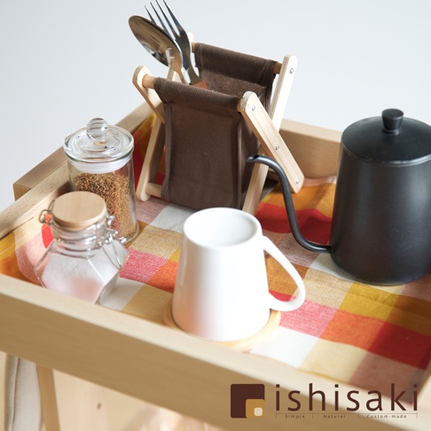木質收納推車上擺放著一個白色的馬克杯、一個黑色細嘴茶壺、兩個玻璃罐（分別裝有淺褐色顆粒和白色粉末）、一個展示餐具（湯匙和叉子）的木質帆布架，以及一塊橘黃色格紋布。