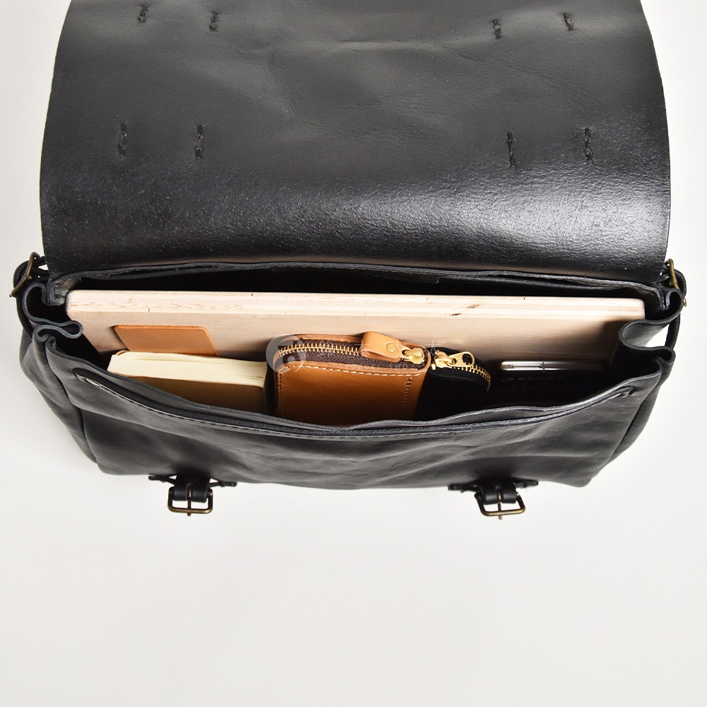 黑色皮革復古郵差包，內部裝有筆記本、一個棕色皮革零錢包、手機和一本薄書。