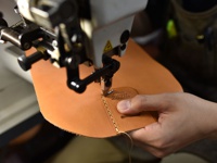 職人手作皮革運動斜背包的製作過程，展示縫紉機正在縫製淺棕色皮革部件，皮革上印有「HERZ」字樣。