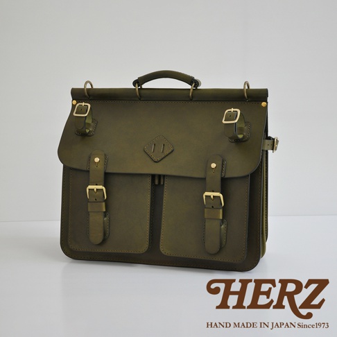 橄欖綠皮革旅行包，配有黃銅色搭扣和兩個前置翻蓋口袋。包的頂部有提把和可調節肩帶的環。