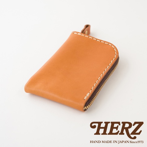 一個來自日本HERZ品牌的棕色職人手作皮革迷你零錢包，採L型拉鍊開口與簡約縫線設計，表面具有皮革自然光澤，右下角配有HERZ品牌商標與產地說明，整體呈現精緻質感。