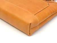 職人手作皮革威靈頓直式拉鍊托特包，採用天然皮革材質，呈現溫潤的焦糖棕色調。包體邊緣以白色縫線精細車縫，展現細膩工藝。包款一角特寫，露出部分拉鍊細節。