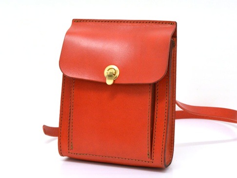 橘紅色厚實皮革製成的手提單肩包，有翻蓋式封口及金色旋轉鎖扣，包身縫線清晰，附有可調節的皮革背帶。