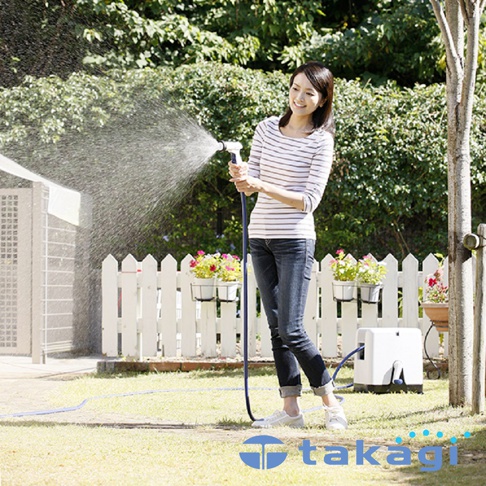 一位女性在庭院中持藍色水管，噴灑水霧，背景有白色栅欄、盆栽花卉及綠色樹叢。一旁有白色捲軸式灑水器。