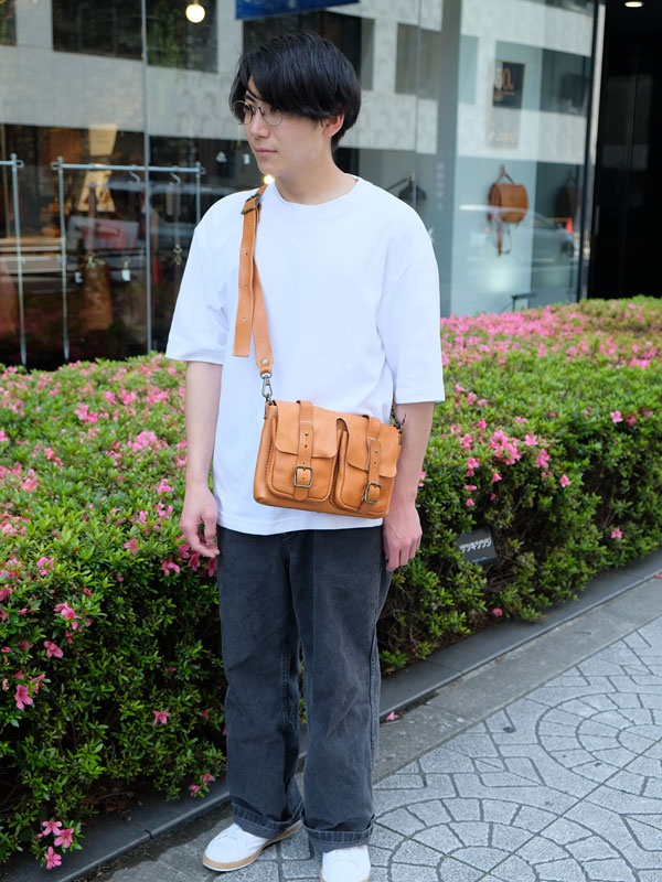 一位男士身穿白色T恤、灰色長褲，斜挎一個棕褐色皮革製雙口袋肩背包，背包上有兩個翻蓋口袋和金屬扣環。