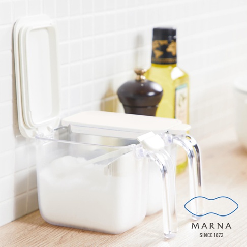 放在淺木紋桌面上的透明塑膠調味料盒，其白色上蓋呈現開啟狀態，露出內部的白色粉末，右側設有透明把手，背景為白色磁磚牆與模糊的廚房用品，右下角印有 MARNA 品牌標誌與成立年份字樣。