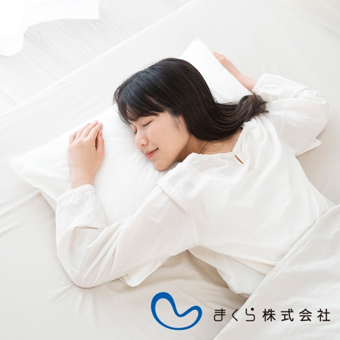 一位女子側臥於白色床鋪，頭枕著白色枕頭，臉上帶著微笑。女子穿著白色長袖上衣，頭髮為黑色。圖片下方有藍色曲線標誌及日文文字。