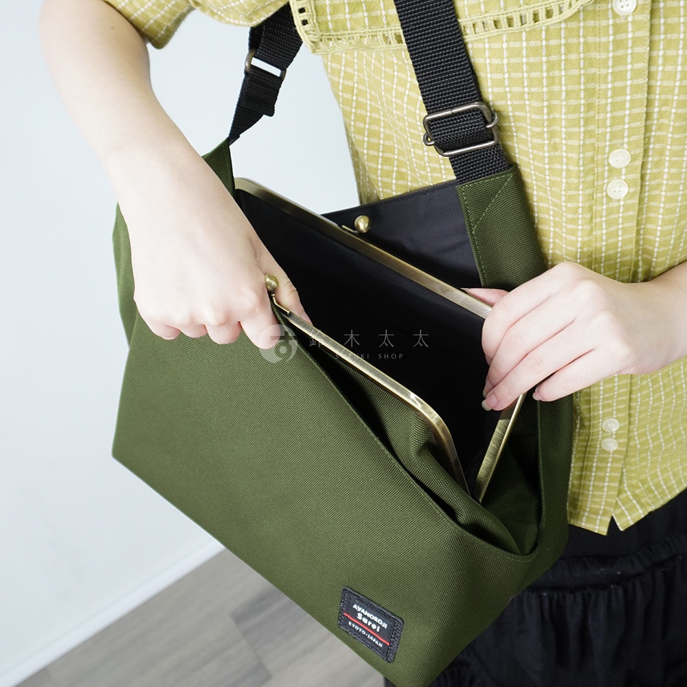 手提著綠色斜背包，包包的金色金屬扣環打開，露出內部黑色內襯，包包底部有 AYANOKOJI Sarei KYOTO-JAPAN 的標籤