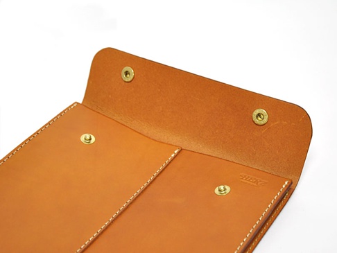 職人手作皮革信封式雙釦手拿袋，展示袋蓋與袋面，採用棕色皮革材質，設有兩個金色金屬按釦，搭配白色車縫線。
