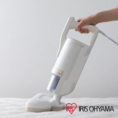 一只手正握著白色塑膠材質的除螨機手把，機器主體為白色，配有透明集塵筒。除螨機的吸頭輕觸在白色布料表面。