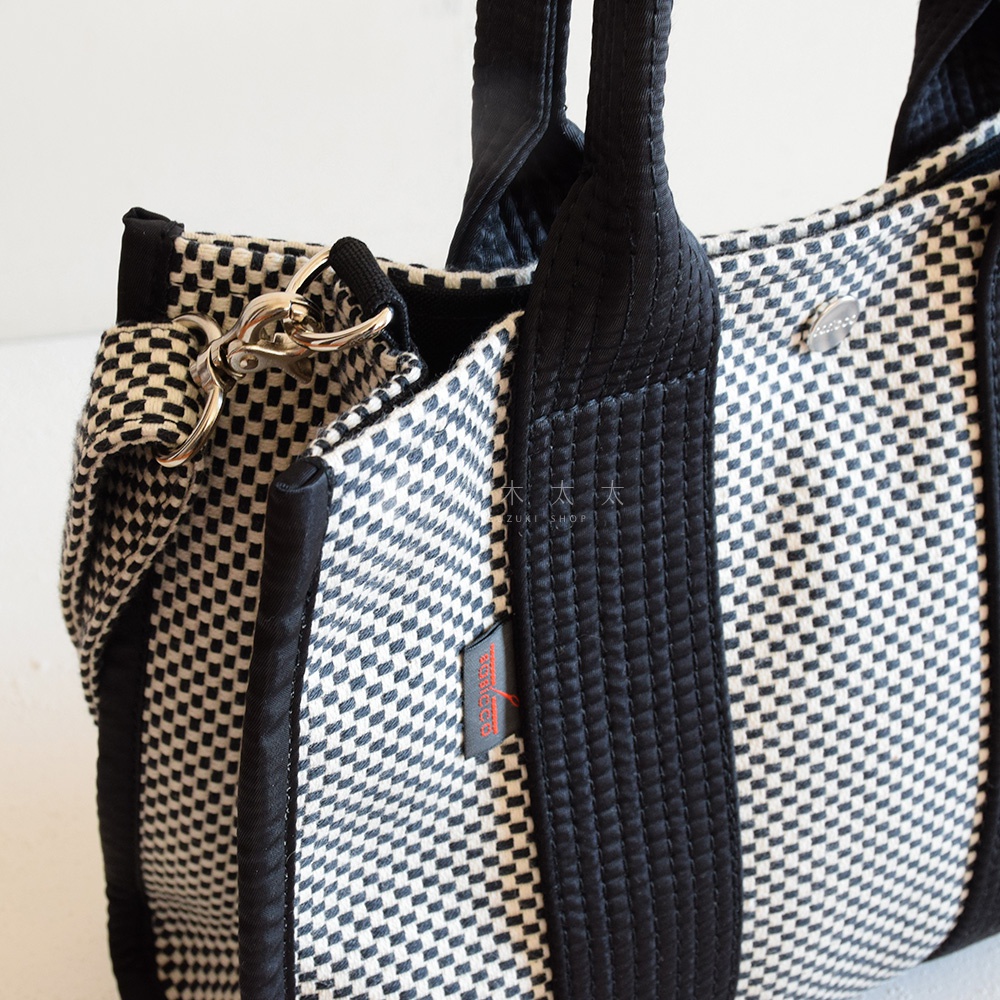 三河木綿刺子織兩用方形托特包，以黑白格紋圖案為主，搭配黑色織帶提把與側面飾條，設有金屬扣環和可拆卸背帶。