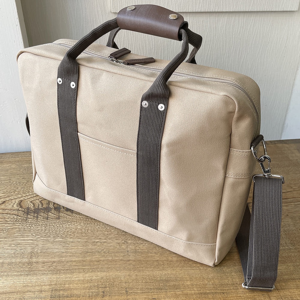 米色純棉帆布公事包，配有深棕色皮革提把和織帶，側面有可拆卸的肩帶，正面有兩個織帶條和一個平口袋，採用銀色金屬拉鍊和鉚釘。