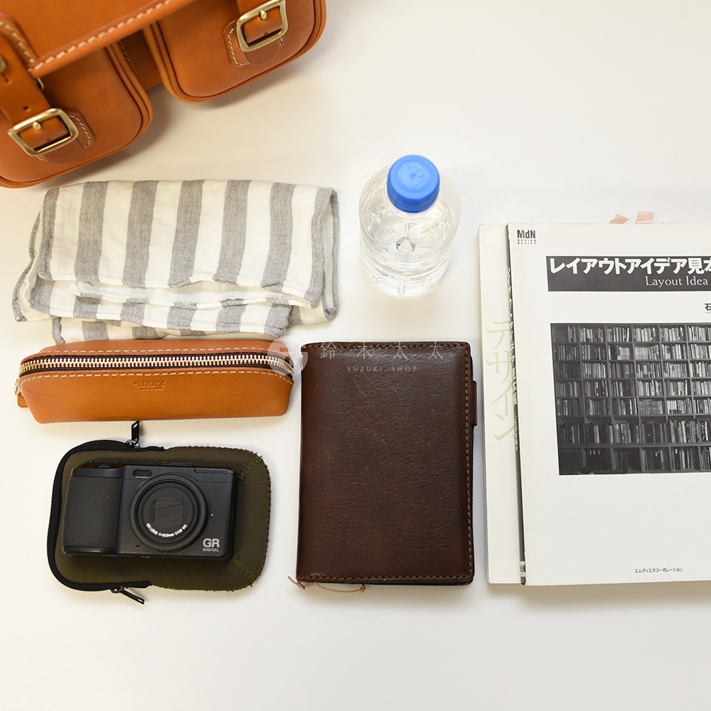 以高角度拍攝，展示一個職人手作皮革經典雙扣郵差包，內含一條條紋毛巾、一個棕色皮革筆袋、一個數位相機、一個深棕色皮夾以及一本日文書籍。