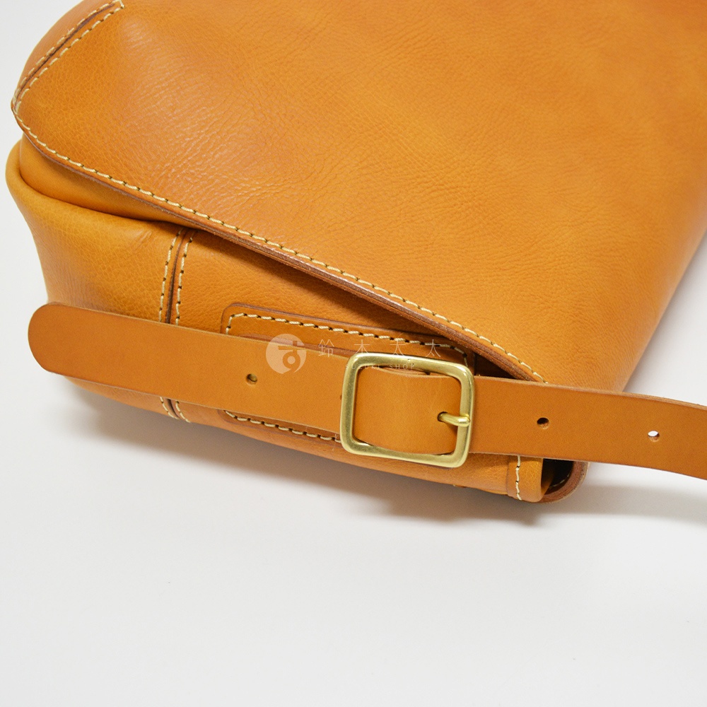 職人手作皮革復古翻蓋郵差包，展示了淺棕色皮革材質，帶有黃銅色金屬釦環和縫線細節。