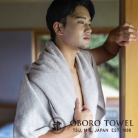 一位年輕男性身披淺灰色細纖浴巾，浴巾質感柔軟，呈現摩登復古風格，搭配品牌 Logo。