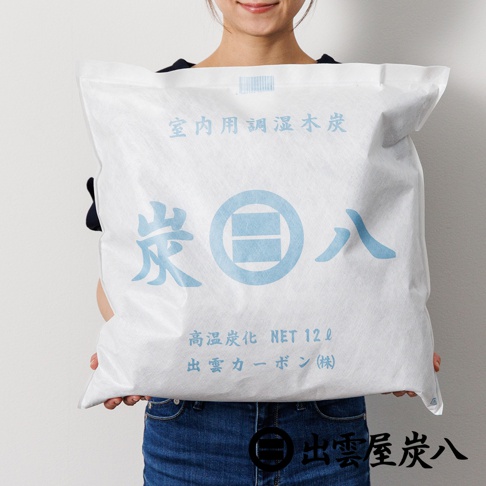 一位女性雙手捧著一個方形白色包裝袋，袋身印有藍色的日文文字與標誌，這是出雲屋炭八的大型室內調濕木炭，包裝正面標示內容物為十二公升的高溫炭化木炭。