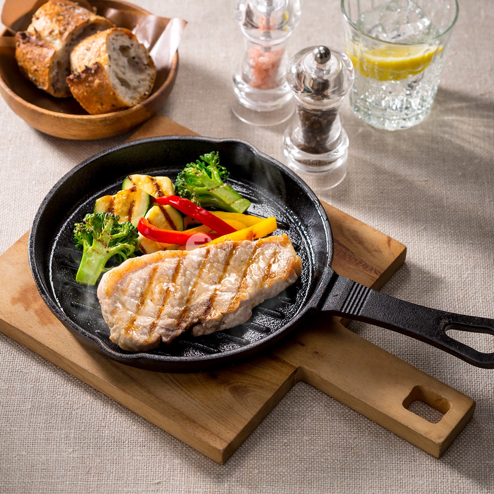 黑色的鑄鐵波浪平底鍋中，盛著烤過的豬排、綠花椰菜、櫛瓜片、紅黃甜椒，鍋柄帶有紋路，平底鍋放置於木製砧板上，周圍有麵包、鹽罐、胡椒罐和一杯檸檬水。