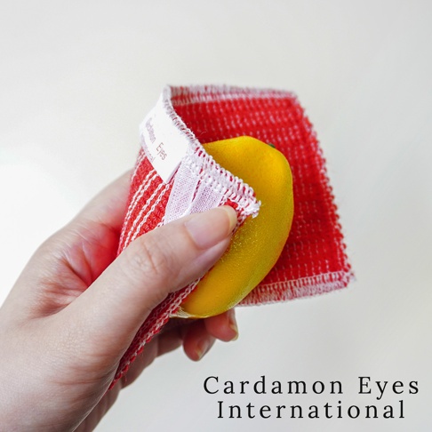 手持紅色條紋壓克力纖維清潔巾，巾上壓有白色品牌標籤，一塊黃色物體（疑似檸檬）被清潔巾包裹。