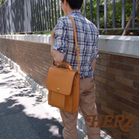 復古風格的棕色皮革手提/斜肩後背包，具有翻蓋開合與金屬鎖扣，側邊有縫線細節，由男性身著格子襯衫與卡其褲展示。