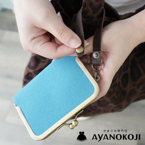 AYANOKOJI 品牌手作帆布橫式口金卡片收納包，藍色帆布搭配古銅色口金，由雙手從皮質掛繩旁取出。