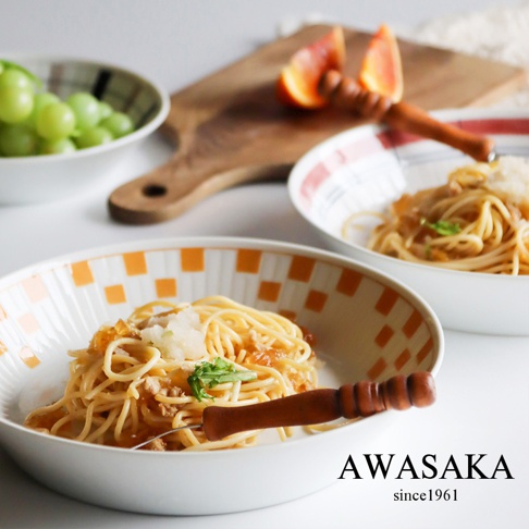 兩個白色陶瓷主食料理盤，其中一個裝有義大利麵和少量配料，盤緣飾有橘色格子圖案。另一盤也盛有義大利麵，盤緣飾有紅色條紋。背景為木製砧板和一碗綠色葡萄。