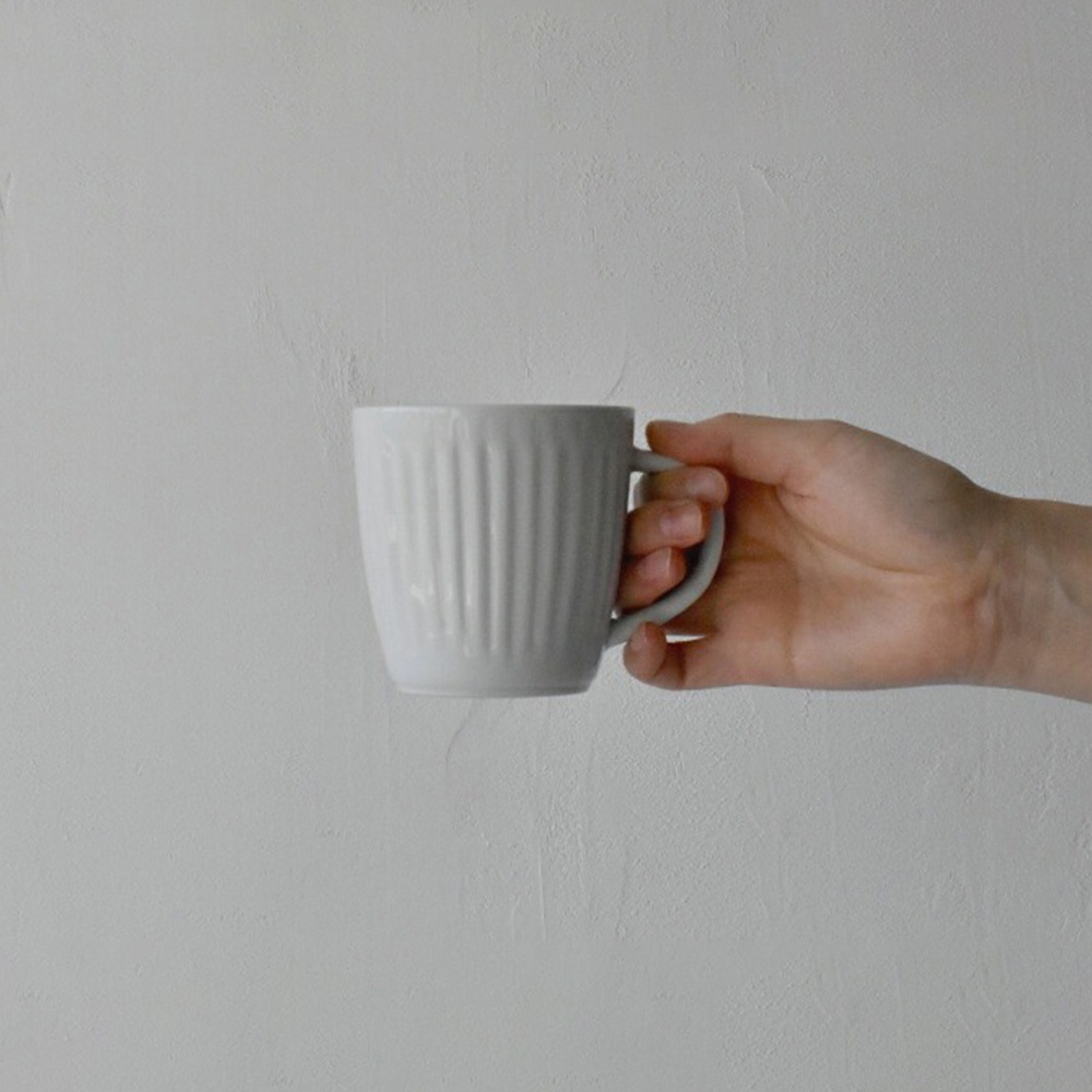 手持一個淺灰色的陶瓷馬克杯，杯身帶有垂直凹槽設計，杯柄呈弧形。