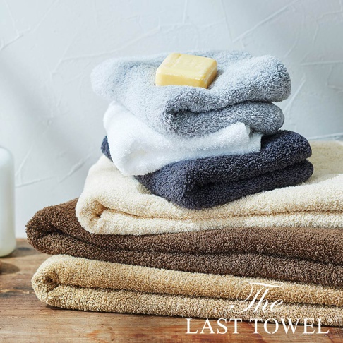 堆疊的純棉長毛巾，包含淺灰色、白色、深灰色、米白色、棕色和淺棕色，頂部有一塊黃色肥皂。