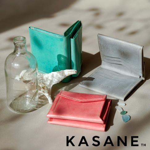 北海道KASANE 品牌字樣，旁邊是三款皮革短夾，分別為綠色、灰色及粉紅色，另有一玻璃瓶和幾顆海玻璃飾品。