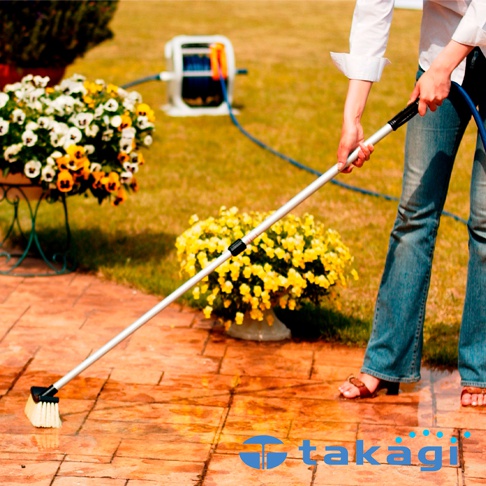 一隻手拿著帶有白色刷毛的銀色伸縮桿，連接到藍色水管，正在清潔磚砌的露台。背景有花卉盆栽和灑水器。