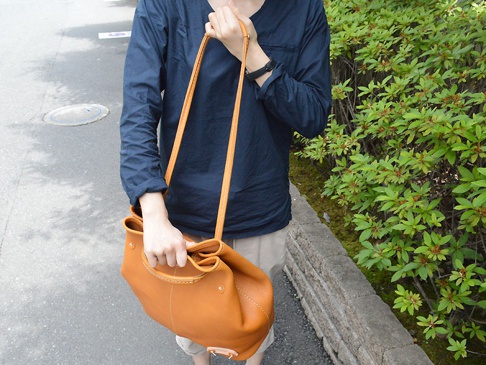 一個棕褐色皮革托特包，配有可調式肩帶，由一人在戶外手持，背景是綠葉植物和鋪砌的道路。