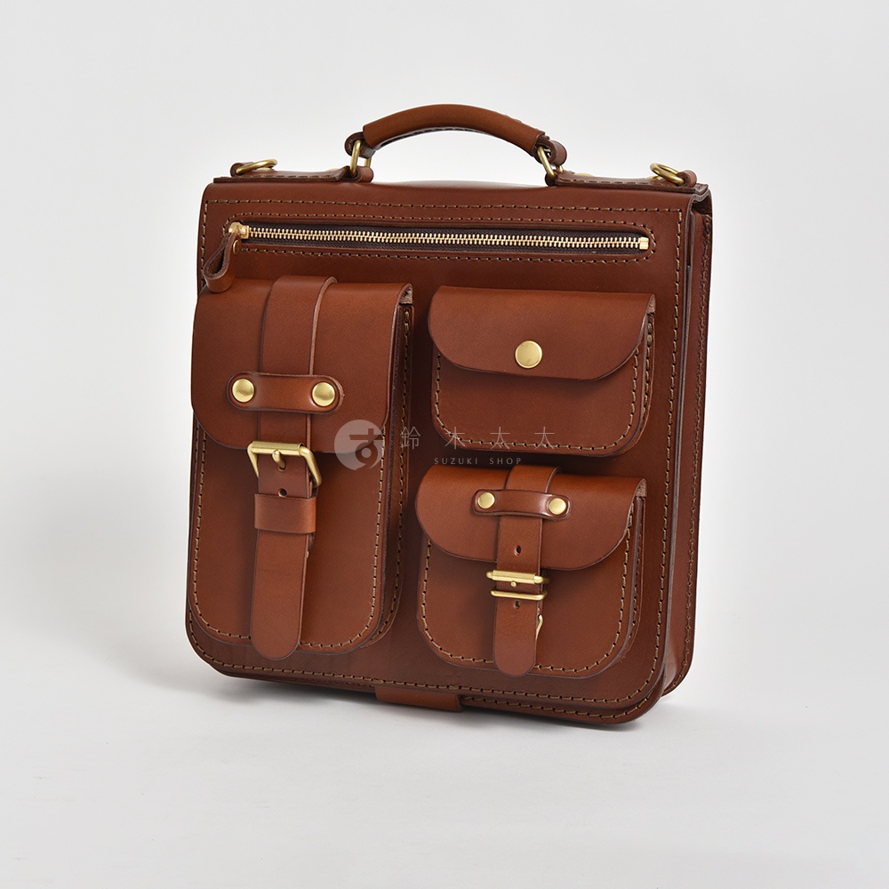 職人手作皮革三用後背包，採用棕色皮革製作，設有三個前置口袋，其中兩個附有皮帶釦和金屬扣環，一個附有金屬鈕扣。包款頂部有拉鍊開口和提把，側面有可拆卸的肩帶固定環。