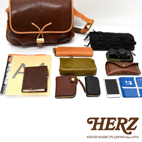 職人手作皮革鹿角扣繩郵差包，包含一個棕色皮革郵差包，以及散落的棕色、橘色、橄欖綠色和黑色皮革配件，如筆記本、零錢包、卡夾、手機，還有一個黑色布質小物袋和藍色印花手帕。