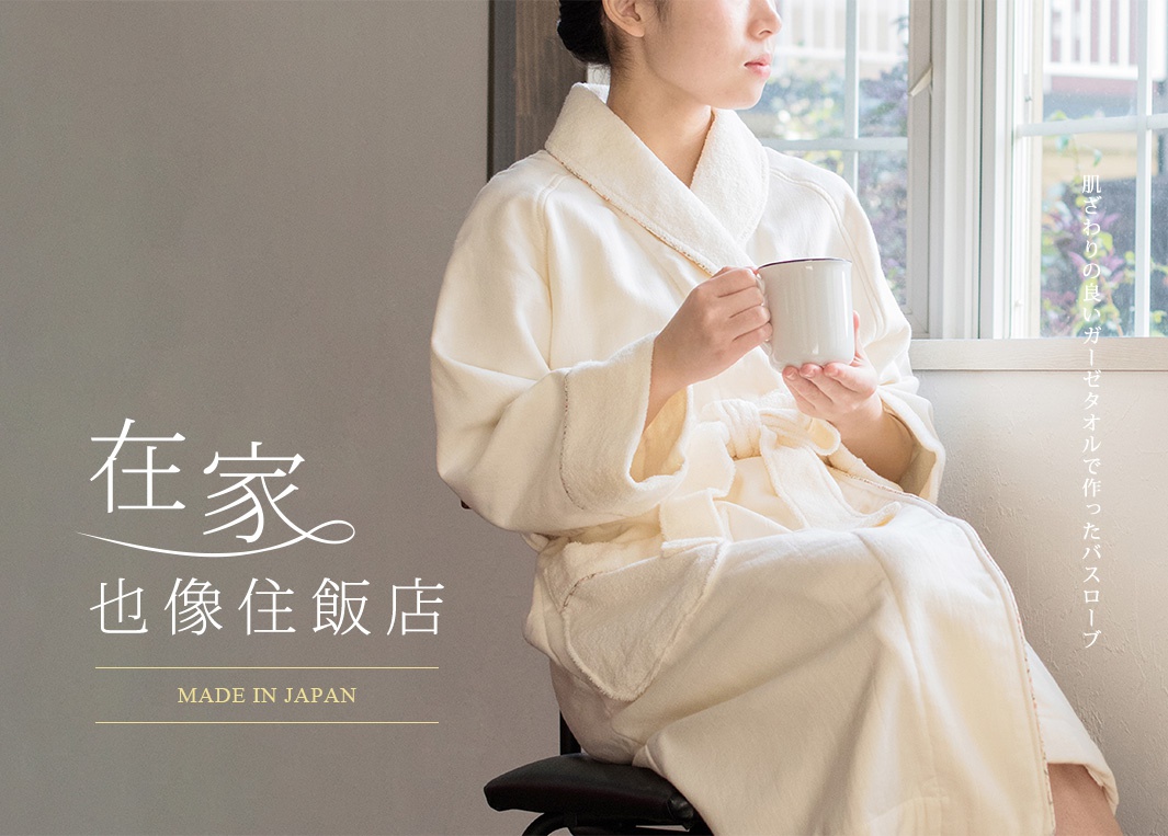 一位女性穿著米白色今治純棉浴袍，手持白色馬克杯，坐在椅子上，靠窗而坐。浴袍材質柔軟，領口和袖口有細緻的縫線。