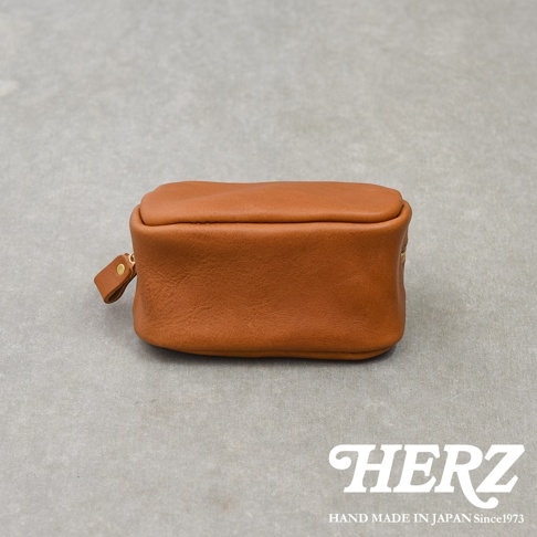 這是一款由日本HERZ品牌職人手作的棕色皮革收納包，呈現長方體造型，表面為自然的皮革紋理，側面配有簡約的拉鍊拉環，整件作品擺放在灰色水泥背景上，右下角印有HERZ品牌標誌與手工製作資訊。