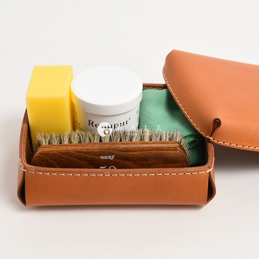 職人手作皮革小物收納盒，內含黃色海綿、白色乳霜罐、綠色拭布及木柄鬃毛刷，整體以淺棕色皮革製成，縫線清晰。