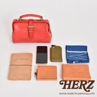 紅色皮革迷你醫生包，附有金色搭扣和手提把。周圍展示一個手機、一個綠色皮革卡夾、一本棕色筆記本、一個橘色皮革短夾、一個橘色皮革化妝包，以及一條藍色印花手帕。