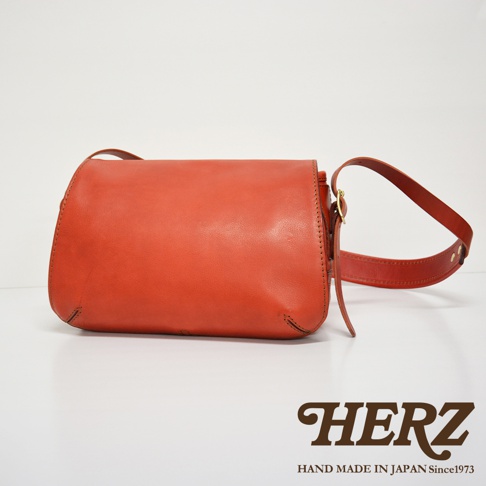 一個橘紅色皮革復古翻蓋郵差包，有可調節的皮質肩帶，包身縫線清晰可見。