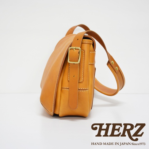 職人手作皮革復古翻蓋郵差包，採用淺棕色皮革，配有金色搭扣和可調節肩帶。包身線條簡潔，車線明顯，展現手工質感。