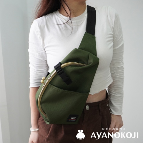 一名女子穿著白色長袖上衣和棕色長褲，斜背一個綠色機能布水滴型口金斜背包。