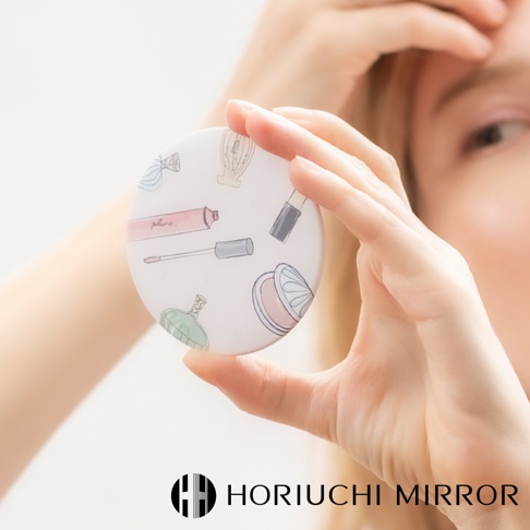 一個圓形隨身小圓鏡，鏡面印有粉紅色唇膏、睫毛膏、口紅、香水瓶及化妝品盒的插畫圖案，背景為白色。