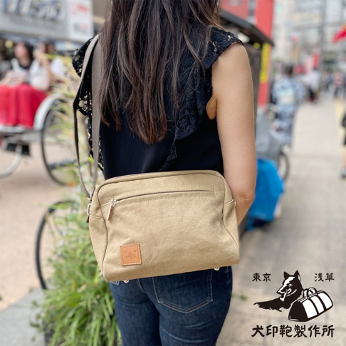 一位女士身著黑色蕾絲上衣和牛仔褲，肩上斜挎著一個石洗帆布旅行側背包。背景是模糊的街道景觀，有日式風格的建築和人物。