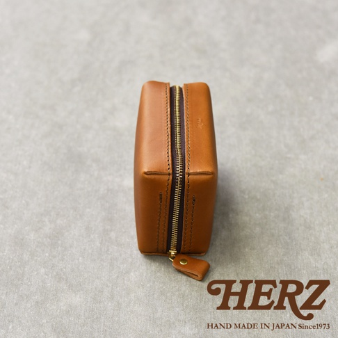 一個方形的棕褐色皮革收納包，帶有黃銅拉鍊，放在灰色水泥背景上。