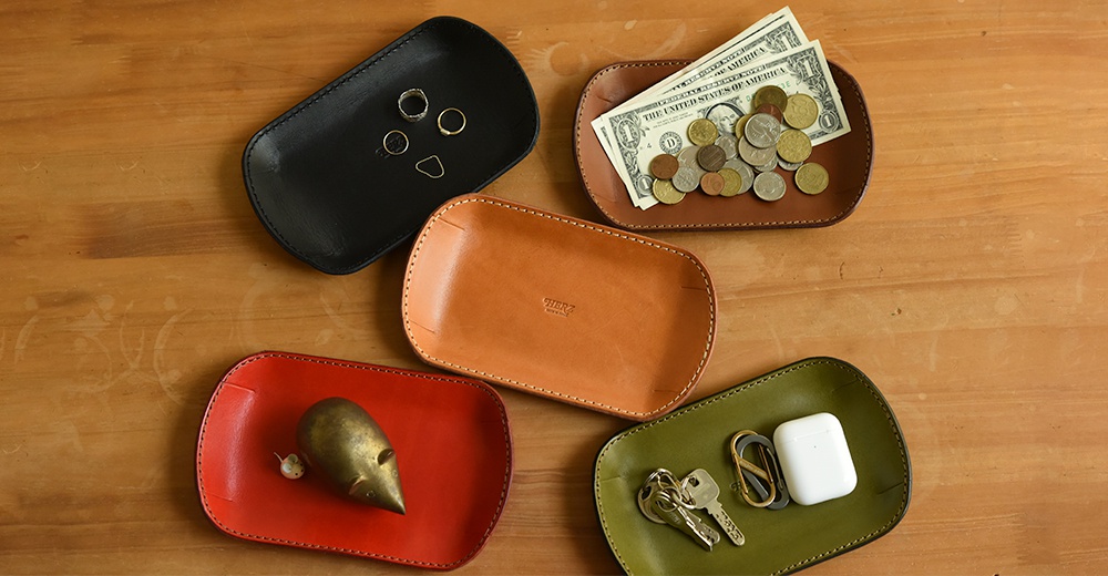 五個職人手作皮革小物收納盤，分別為黑色、棕色、橘色、紅色和綠色。盤子由皮革製成，邊緣有縫線裝飾。黑色盤子內有戒指，棕色盤子內有鈔票和硬幣，紅色盤子內有金屬老鼠擺飾，綠色盤子內有鑰匙和無線耳機。