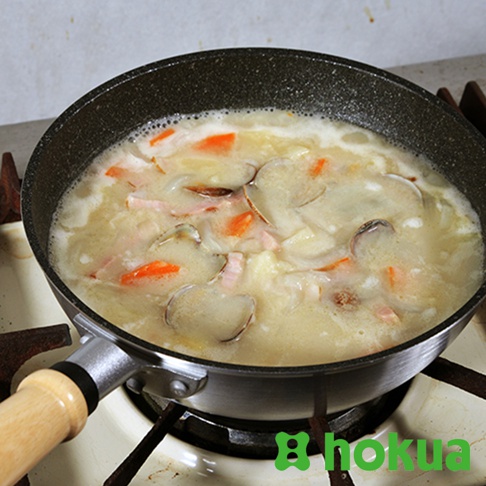 一個黑色炒鍋正在瓦斯爐上烹煮湯品，鍋內有蛤蜊、紅蘿蔔塊和肉片，鍋柄為淺棕色木質。