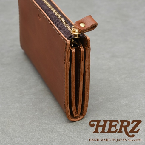 HERZ 品牌標誌，下方為 HAND MADE IN JAPAN Since1973 字樣，圖片主體為一個棕色皮革拉鍊長夾，採側面視角，拉鍊為金色，細節處可見皮革的紋理及縫線。