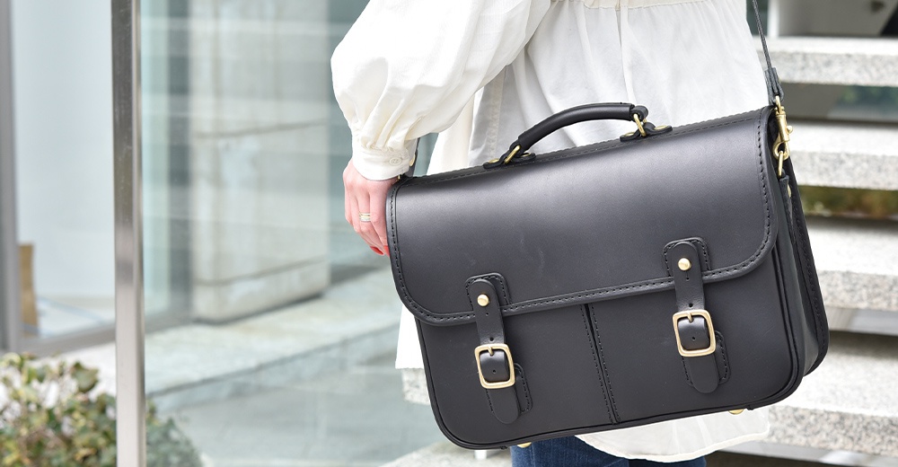 黑色皮革製成的職人手作單肩包，配有頂部提把、可調節肩帶、黃銅色金屬扣環和裝飾釘，適合商務或休閒場合。