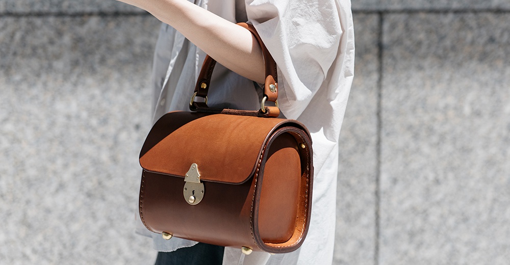 棕色皮革復古手提包，帶有金色鎖扣和提把，側邊有縫線細節，底部有四個金色支腳。